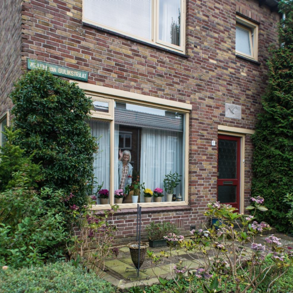Seniore dame zwaait achter het raam vanuit woonhuis (foto Alex Schröder)