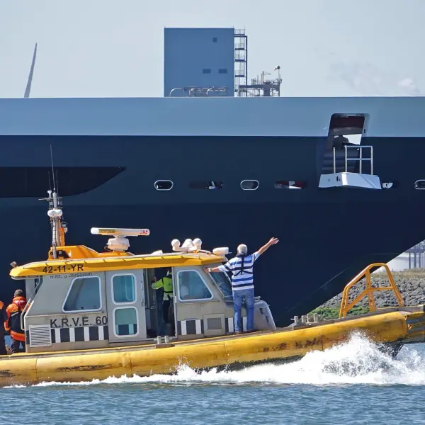 Vier loodsen op een gele boot begeleiden een groot schip door de haven bij Hoek van Holland. Een van de loodsen lijkt met een armgebaar de weg te wijzen.