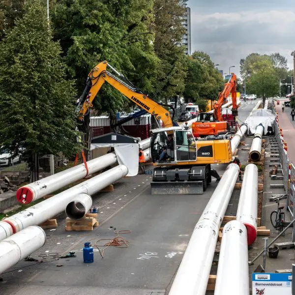 Straat met oude huizen waarin gewerkt wordt. Op de foto zie je - naast bomen, fietsers en voetgangers - grote witte buizen, en een graafmachine.