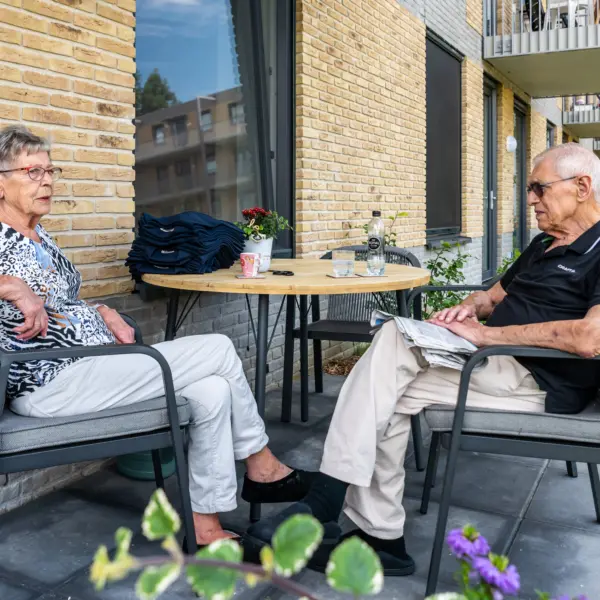 Twee senioren zitten tegenover elkaar op een balkon bij een appartementencomplex en praten samen aan een tafel met koffie en een krant.