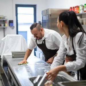 A chef and cook cleaning the workspace after doing dishes indoors in restaurant kitchen.