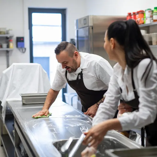 A chef and cook cleaning the workspace after doing dishes indoors in restaurant kitchen.