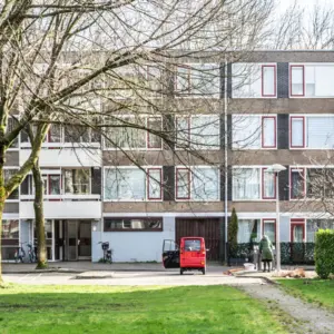 Flat met grasveld en bale bomen, voor de flat staat een rood rolstoel-wagentje. Een vrouw in een warme jas gooit iets in de afvalcontainer naast het wagentje.