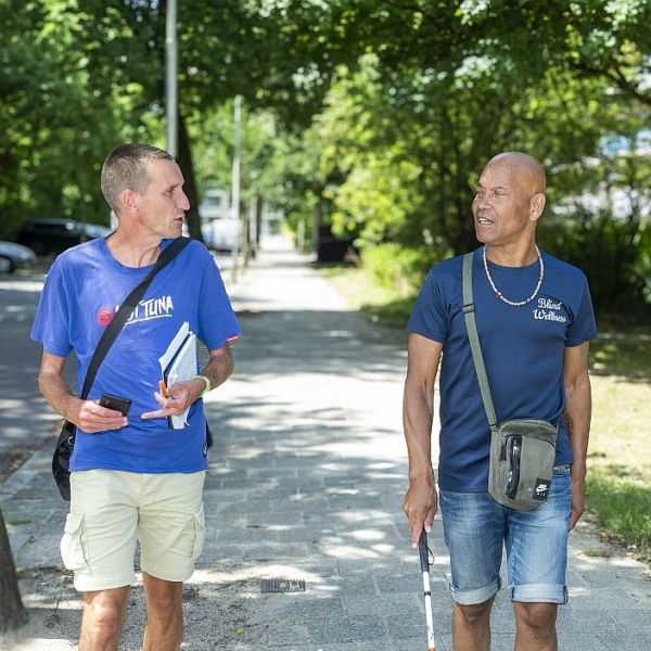 Twee mannen wandelen op straat in de halfschaduw onder bomen. Het is een wandelinterview voor een 15 minute ctiy-onderzoek in Delft